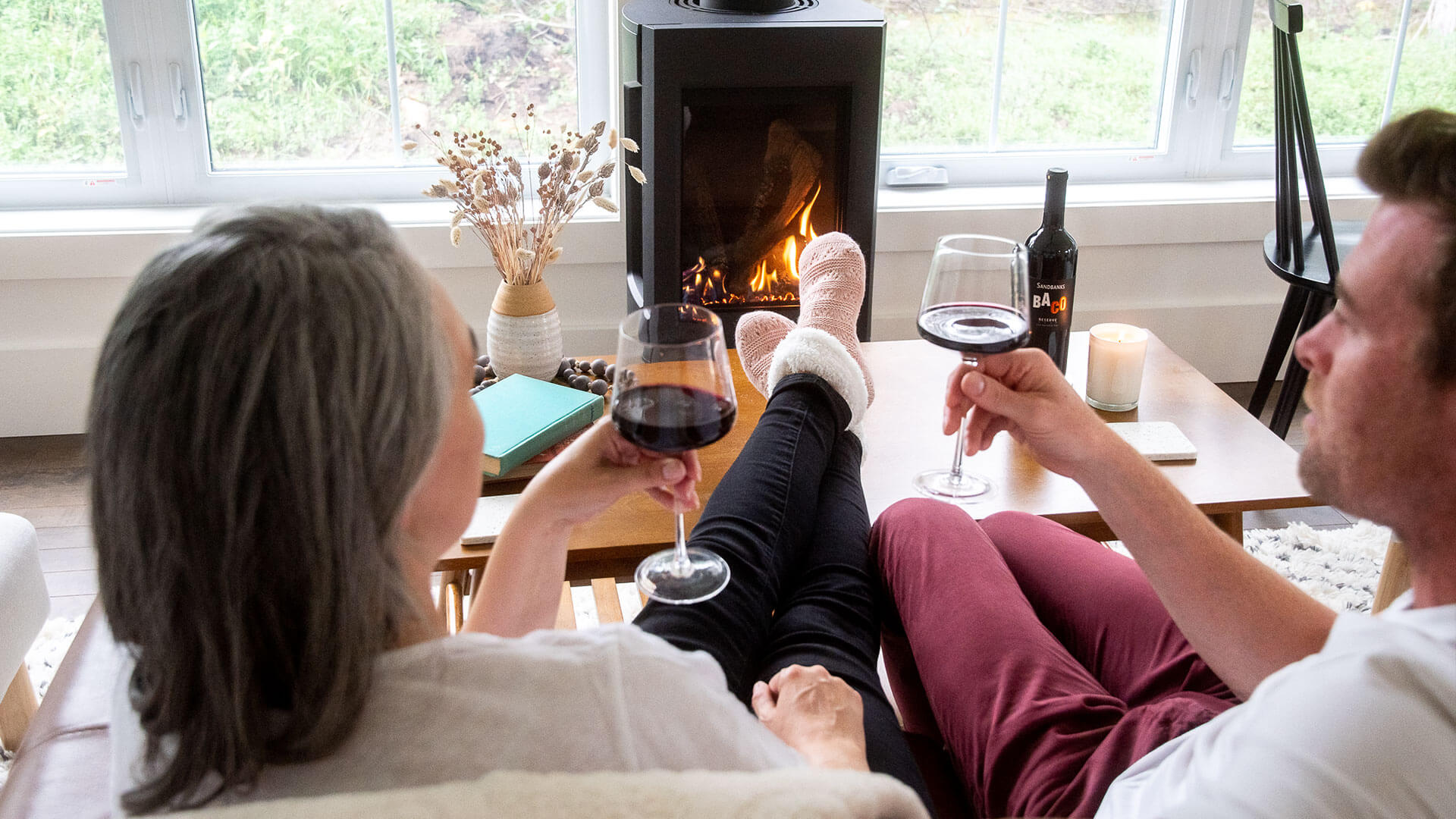 Couple enjoying a glass of Sandbanks Reserve Baco Noir VQA with their feet up by their fireplace.