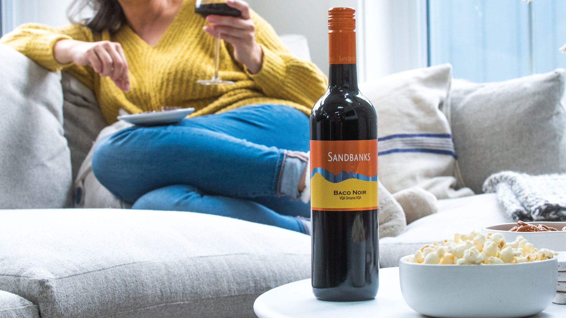 Wine enthusiast enjoying a glass of Sandbanks Baco Noir on the sofa with some popcorn nearby. 