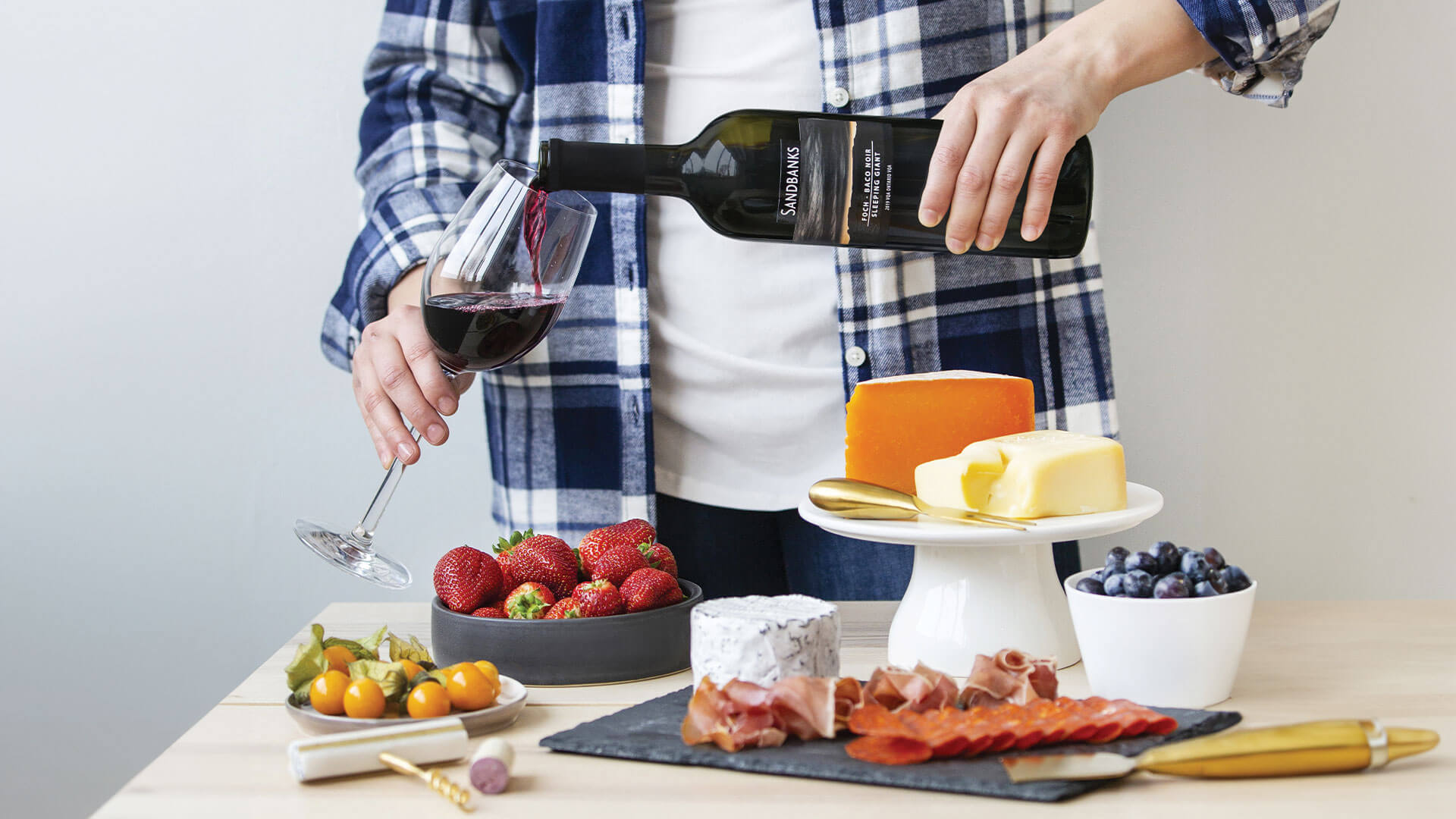 Person pouring a glass of Sandbanks red wine to pair with cheese, cured meat, and berries.  